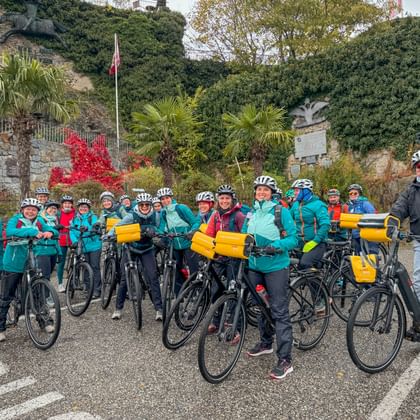 Large group of cyclists in turquoise jackets with helmets and bikes with yellow panniers, posing in front of stone wall with palm trees.