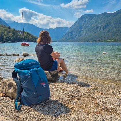Wanderer mit blauem Rucksack sitzt auf Felsen am Ufer des Bohinjer Sees. Türkisfarbenes Wasser, Segelboot und Julische Alpen unter blauem Himmel.
