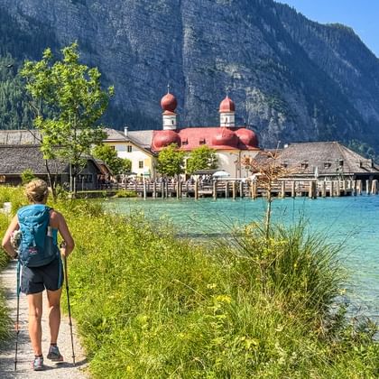Wanderin mit Rucksack auf Uferweg am Königssee. St. Bartholomä Kirche mit roten Kuppeln über türkisfarbenem Wasser, Berge im Hintergrund.