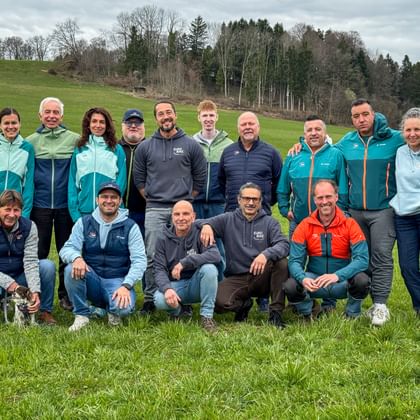 Group photo of 16 people in outdoor clothing on a green meadow. Rolling hills and forest visible in the background under cloudy sky.