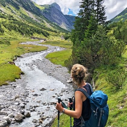 Female hiker with blue backpack and trekking poles on Lechweg, overlooking a green mountain valley with a winding stream.