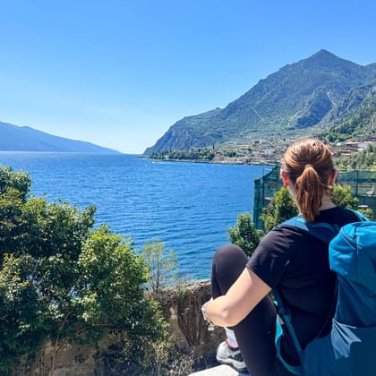 Wanderin mit blauem Rucksack blickt auf den Gardasee und Limone sul Garda, mit Bergen und blauem Wasser unter klarem Himmel.