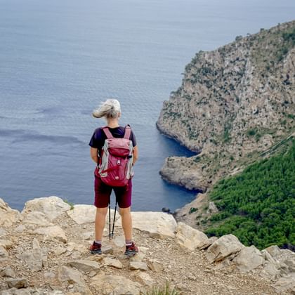 Female hiker with backpack and trekking poles standing on rocky cliff overlooking the Mediterranean Sea near Alcudia, Mallorca.