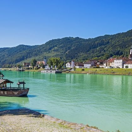 Traditional wooden ferry on turquoise Danube River near Engelhartszell with colorful buildings and forested hills in background under blue sky.