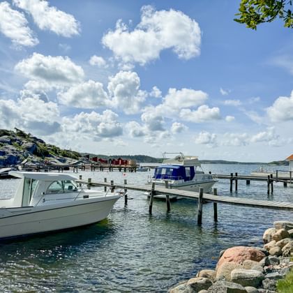 Weiße Motorboote an Holzstegen im Hafen von Särö. Felsige Küste im Vordergrund, Gebäude mit rotem Dach sichtbar, blauer Himmel mit weißen Wolken.