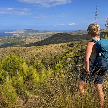 Wanderin mit blauem Rucksack auf Hügel mit Blick auf Mallorcas Ostküste mit türkisfarbenem Meer und bergiger Landschaft.