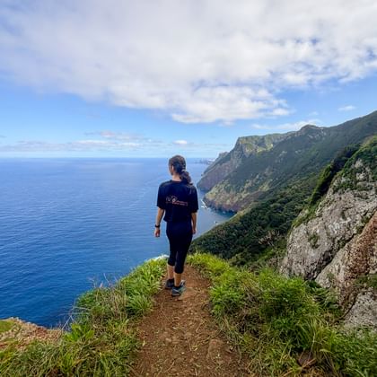 Female hiker walking on Vereda do Larano coastal trail in Madeira with steep cliffs, green vegetation, and deep blue Atlantic Ocean.