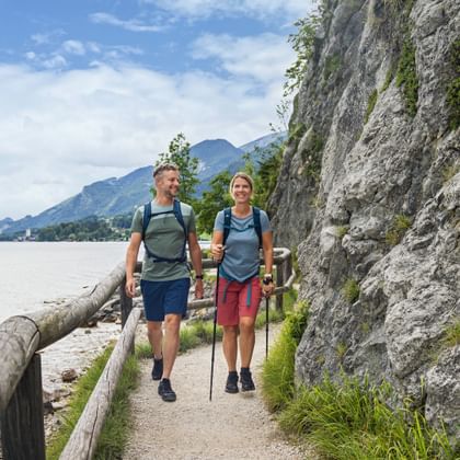 Zwei Wanderer mit Rucksäcken und Wanderstöcken auf einem schmalen Weg zwischen Felswand und See, mit Bergen im Hintergrund.