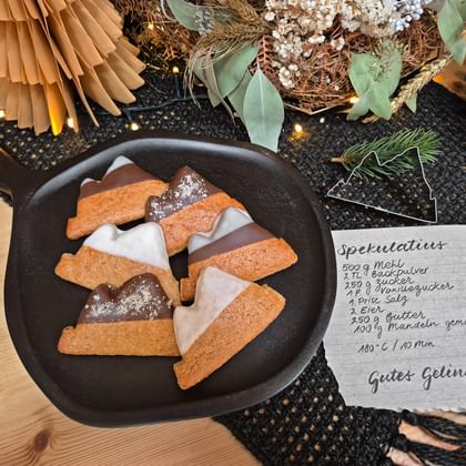 Cast iron pan with glazed speculatius cookies beside a handwritten recipe card, surrounded by festive decorations including paper fans and greenery.