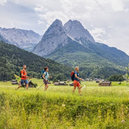Drei Wanderer mit Rucksäcken gehen durch grüne Wiesen mit dramatischen Alpengipfeln im Hintergrund bei Garmisch-Partenkirchen.
