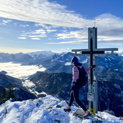 Wanderer in lila Jacke steht am Gipfelkreuz auf schneebedecktem Gipfel mit Blick auf verschneite Alpentäler und Bergketten unter blauem Himmel.
