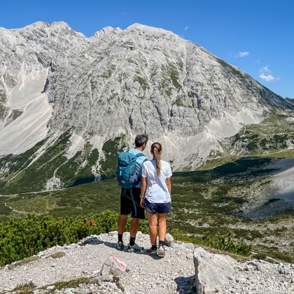 Two hikers standing on rocky viewpoint overlooking Karwendel mountain range with dramatic limestone peaks and green alpine valleys below.
