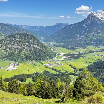 Alpine valley view from Buchensteinwand near Kitzbühel with green meadows, forested hills, small villages, and snow-capped mountains under blue sky.