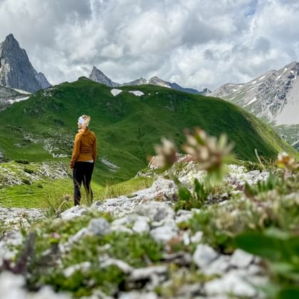 Wanderin in oranger Jacke auf Almwiese mit Wildblumen, blickt auf die Weissschrofenspitze und dramatische Berglandschaft.