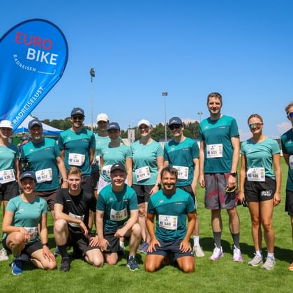 Group of runners in turquoise shirts with race bibs posing on grass field. Eurobike flag visible on left under clear blue sky.