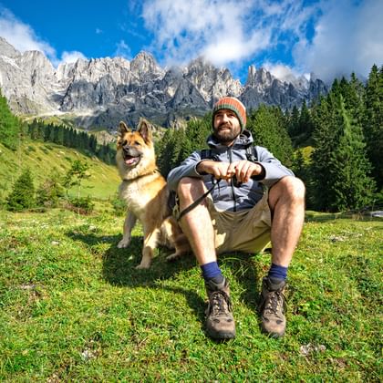 Mann in Wanderausrüstung sitzt auf Almwiese mit braunem Hund neben sich. Felsige Berggipfel und grüne Wälder im Hintergrund unter blauem Himmel.