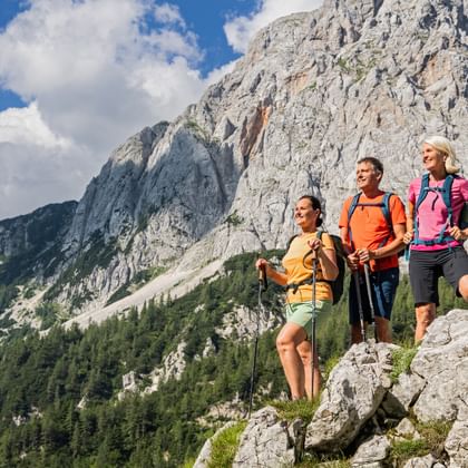 Three hikers with backpacks standing on rocky terrain at Vrsic Pass, with dramatic limestone peaks and forested slopes under blue sky.