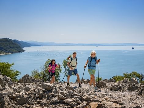 Drei Wanderer mit Rucksäcken und Wanderstöcken stehen auf felsigem Gelände mit Blick auf die Adria bei Triest in den Julischen Alpen.