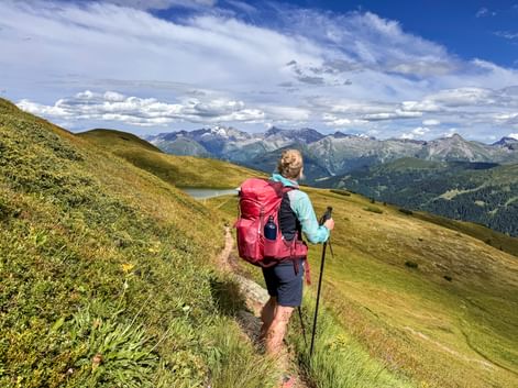 Hiker with red backpack walking on mountain trail near Obernberg with panoramic view of Alpine peaks and green slopes under blue sky.