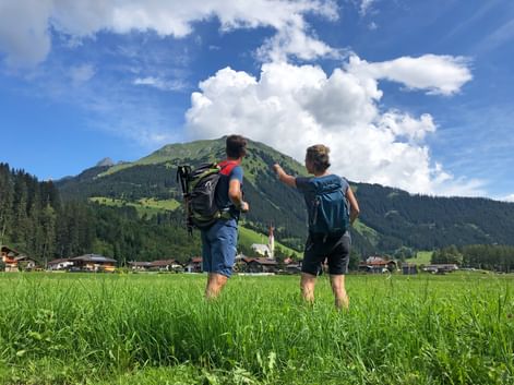 Zwei Wanderer mit Rucksäcken stehen auf einer grünen Wiese am Lechweg und zeigen auf einen Berg. Alpendorf und Kirche unter Bergen sichtbar.