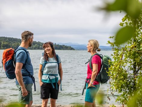 Drei Wanderer mit Rucksäcken stehen am Seeufer, umgeben von grünem Laub. Berge und bewaldete Hügel im Hintergrund sichtbar.