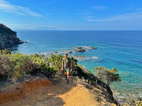 Wanderer mit Rucksack auf Küstenpfad in Cinque Terre mit türkisfarbenem Mittelmeer, felsigen Klippen und grüner Vegetation.