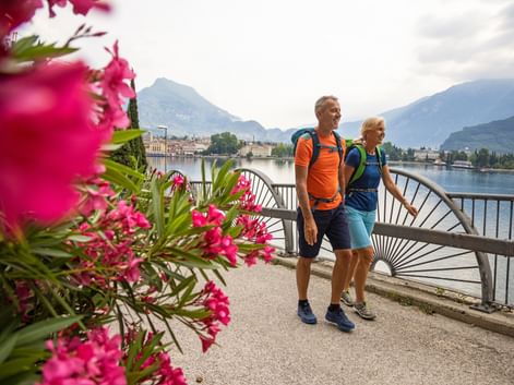 Two hikers with backpacks standing on a lakeside promenade at Lake Garda, with pink flowers in foreground and mountains in background.