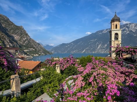 Church tower with pink flowers in Limone sul Garda overlooking Lake Garda, surrounded by mountains under blue sky.