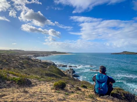 Wanderer mit blauem Rucksack sitzt auf Küstenklippe mit Blick auf türkisfarbenen Atlantik am Praia do Sissal, mit Sandstrand und felsiger Küste.
