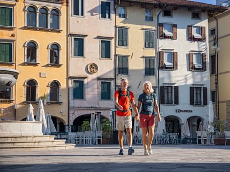 Zwei Wanderer gehen Hand in Hand über einen historischen Platz in Udine, Friaul. Bunte italienische Gebäude mit grünen Fensterläden säumen die Piazza.
