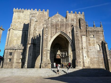 Kathedrale von Tui mit befestigten Steintürmen und gotischem Eingangsportal. Besucher steigen breite Steintreppen zum Hauptportal unter blauem Himmel.