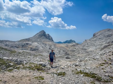 Hiker walking on rocky terrain in the Julian Alps, Slovenia. Gray limestone peaks rise under blue sky with white clouds.