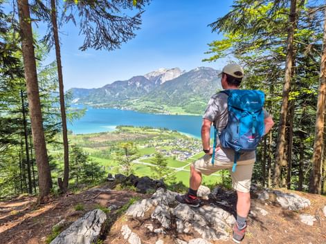 Hiker with blue backpack standing on rocky viewpoint overlooking Wolfgangsee lake in Salzkammergut, surrounded by forest and mountains.