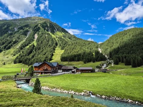 Almhütten des Krimmler Tauernhauses in grünem Tal mit türkisem Bach, umgeben von bewaldeten Bergen unter blauem Himmel in den Hohen Tauern.