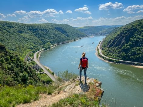 Hiker with red backpack standing on cliff edge overlooking Rhine River between Mainz and Koblenz, with forested hills and boats on the water.