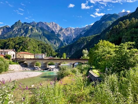 Steinbrücke über türkisfarbenen Fluss im Tal der Julischen Alpen mit traditionellen Gebäuden und dramatischen Berggipfeln unter blauem Himmel.