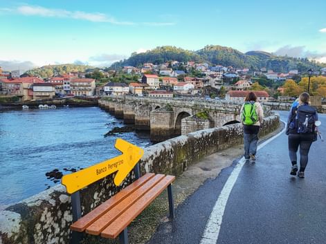 Zwei Wanderer mit Rucksäcken auf der Brücke Ponte Sampaio mit gelbem Camino Peregrino-Schild. Steinbrücke überquert Fluss mit Hügelstadt im Hintergrund.