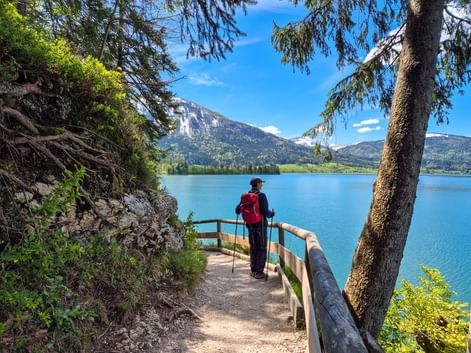 Wanderer mit rotem Rucksack auf Uferweg am Wolfgangsee, Salzkammergut. Holzgeländer säumt Pfad mit blauem See und Bergen im Hintergrund.