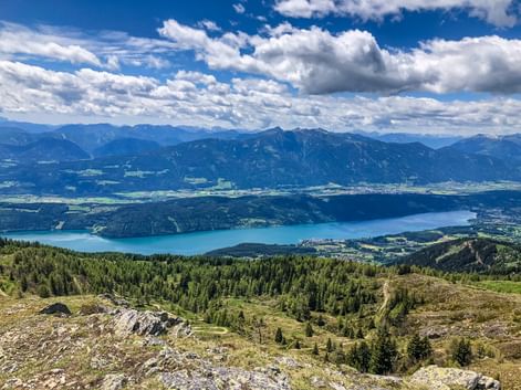 Panoramic view from Gerlitzen mountain showing a large blue lake surrounded by forested hills and distant Alpine peaks under cloudy sky.