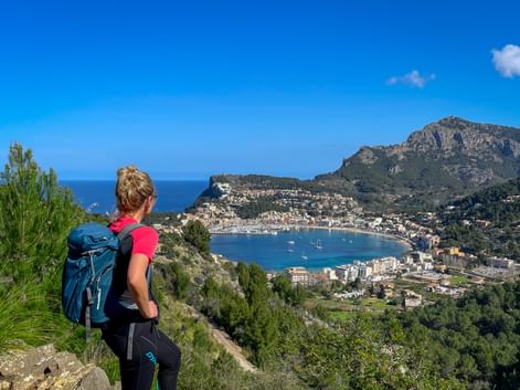 Female hiker with blue backpack viewing Port de Sóller bay from hillside on GR221 trail in Mallorca. Mountains and Mediterranean Sea visible.