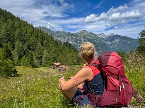 Female hiker with burgundy backpack sitting in alpine meadow near Trins, Gschnitztal, viewing mountain peaks and traditional huts below.