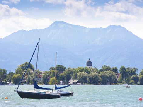 Blaues Segelboot auf dem Chiemsee mit Kirchenkuppel auf bewaldeter Insel und Bergkette im Hintergrund unter teilweise bewölktem Himmel.