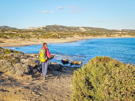 Wanderin mit gelbem Rucksack auf Küstenweg mit Blick auf blaue Bucht mit Sandstrand und Hügeln in Zypern.
