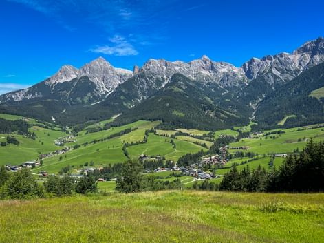 Panoramic view of Maria Alm valley with green meadows, scattered villages, and dramatic rocky mountain peaks under blue sky.