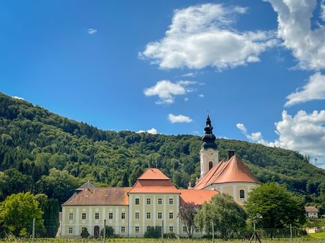 Engelszell Abbey complex with yellow monastery building, church with orange roof and black onion dome tower, set against forested hills.