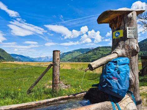 Traditioneller Holzbrunnen mit blauem Wanderrucksack im Pinzgau. Grüne Wiesen und Bergketten unter blauem Himmel mit weißen Wolken im Hintergrund.