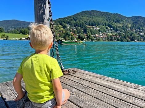 Blondes Kind im gelben Shirt sitzt auf Holzsteg am Tegernsee. Türkisfarbenes Wasser, grüne Berge und Dorf im Hintergrund unter blauem Himmel.