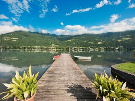 Wooden pier extending into calm Wörthersee lake with boats moored nearby. Green mountains and village reflected in water under blue sky.