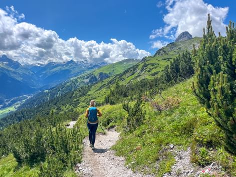 Female hiker with backpack walking on mountain trail through green alpine landscape with snow-capped peaks and blue sky with white clouds.