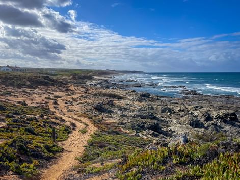 Sandiger Wanderweg entlang felsiger Küste bei Praia do Queimado nahe Porto Covo. Grüne Vegetation säumt den Pfad, dunkle Felsen am Ufer.